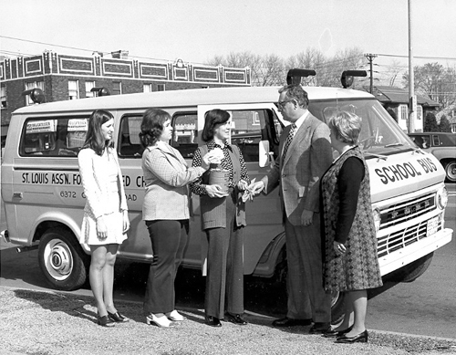 Three women hand coupons to a man while a van sits behind them.