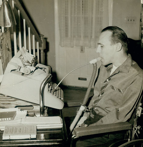 A seated man types on a typewriter using a mouth stick.
