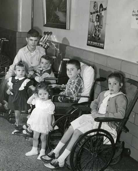 Children of various ages sit in a hallway, several of them in wheelchairs.