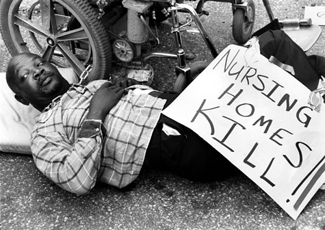 A man lies on the ground next to his wheelchair, holding a sign that says 'Nursing Homes Kill.'