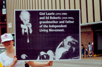 A woman holds up a sign that depicts Gini Laurie and Ed Roberts and includes the caption 'Gini Laurie and Ed Roberts, grandmother and father of the Independent Living Movement.'