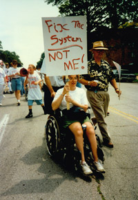 A protester who uses a wheelchair holds up a sign that says 'Fix The System: Not Me.'